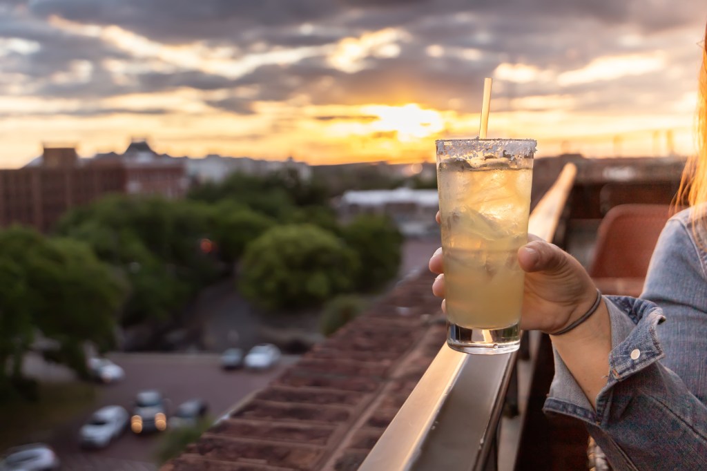 A hand holding a yellow alcoholic beverage with a straw and salted rim at sunset at a rooftop bar, with the tree-lined road beneath out of  focus